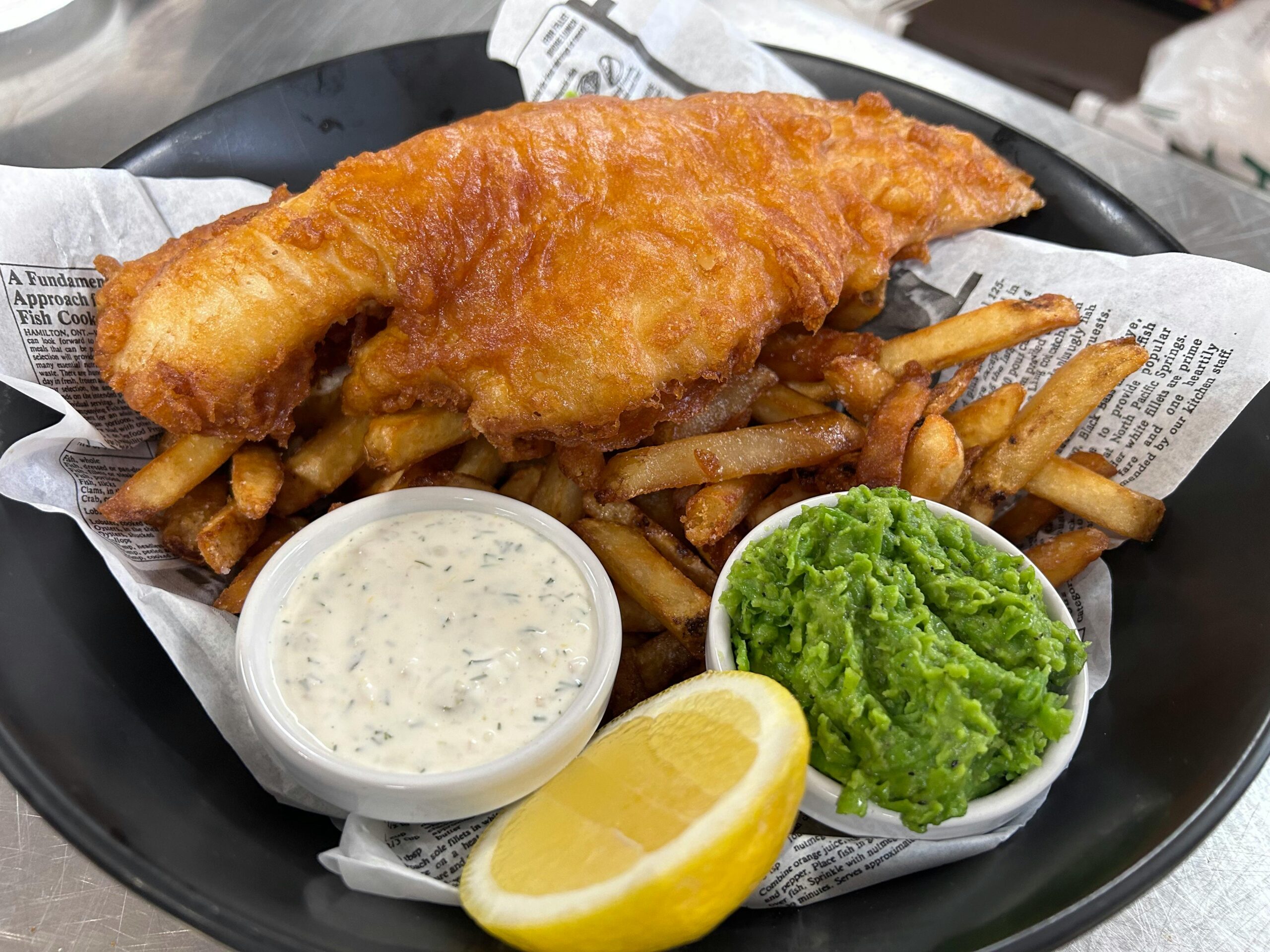 Delicious fish and chips served with tartar sauce, mushy peas, and a lemon wedge in a black dish.